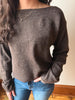 A woman wearing a brown knit sweater with a wide boat neckline and long sleeves, paired with blue jeans, standing indoors against a light-colored wall.