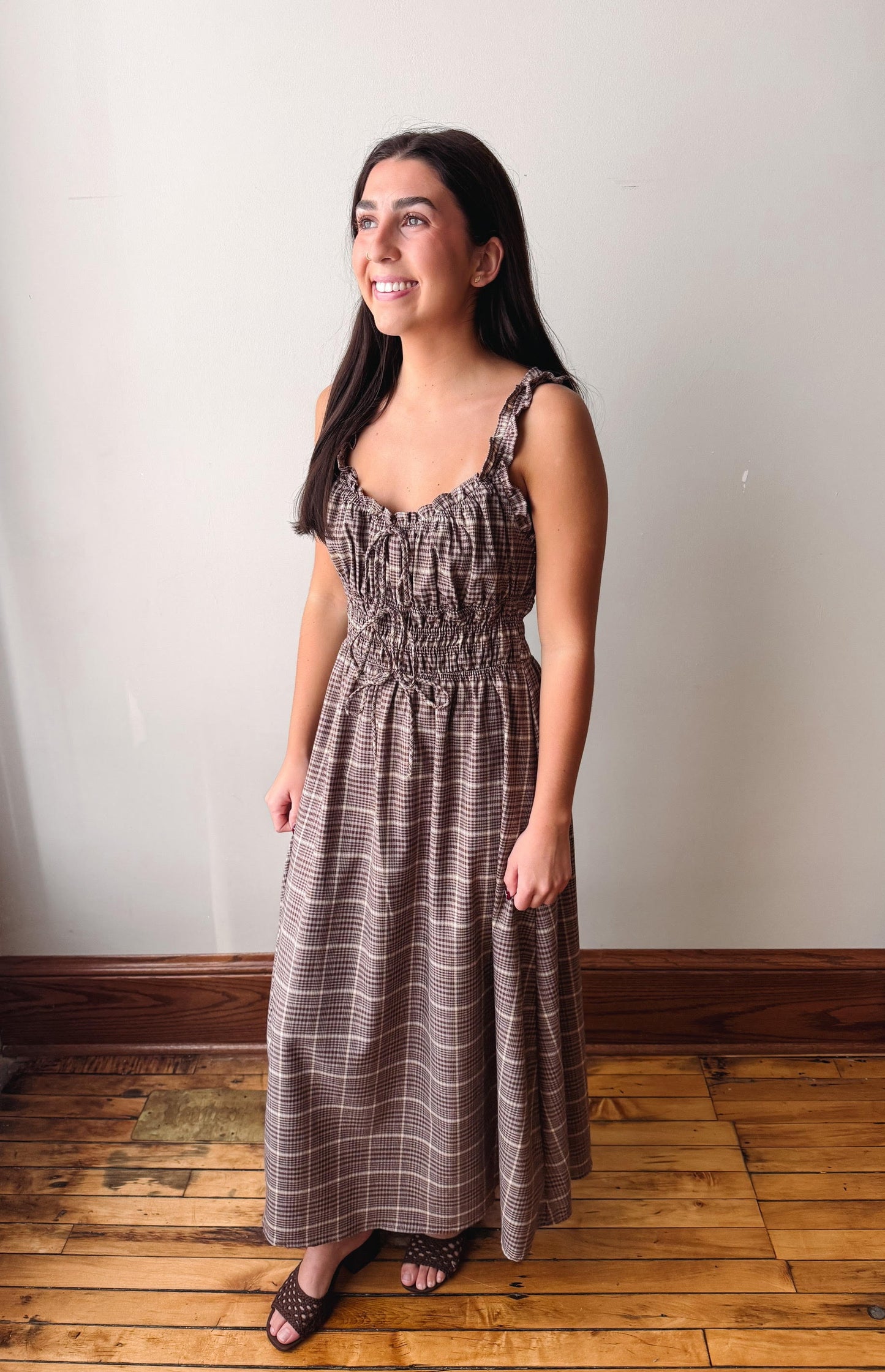 A woman modeling a brown and beige plaid maxi dress with a smocked bodice and spaghetti straps, standing on a wooden floor against a plain off-white wall.