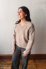 A woman wearing a taupe knit sweater with floral cable patterns and dark gray pants, standing in front of a plain wall with wood flooring.