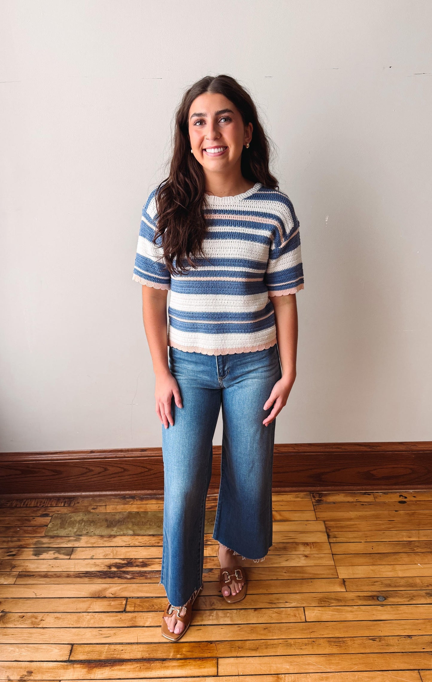 Woman wearing a striped shirt and jeans standing on a wooden floor with a white wall background