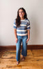 Woman wearing a striped shirt and jeans standing on a wooden floor with a white wall background