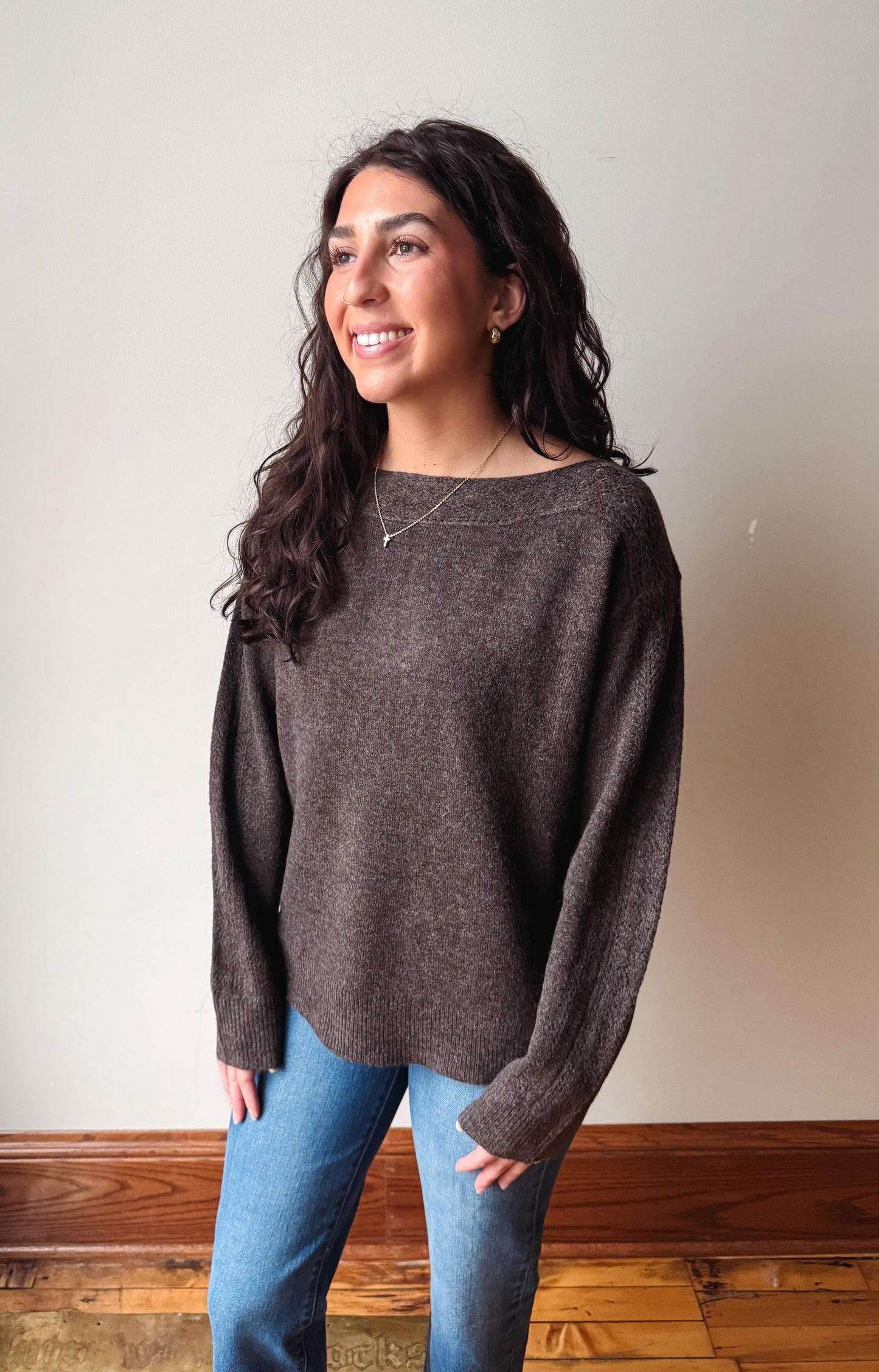 A woman with long dark wavy hair wearing a dark brown knit sweater and blue jeans, standing in front of a plain beige wall with a wooden baseboard.
