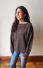A woman with long dark wavy hair wearing a dark brown knit sweater and blue jeans, standing in front of a plain beige wall with a wooden baseboard.