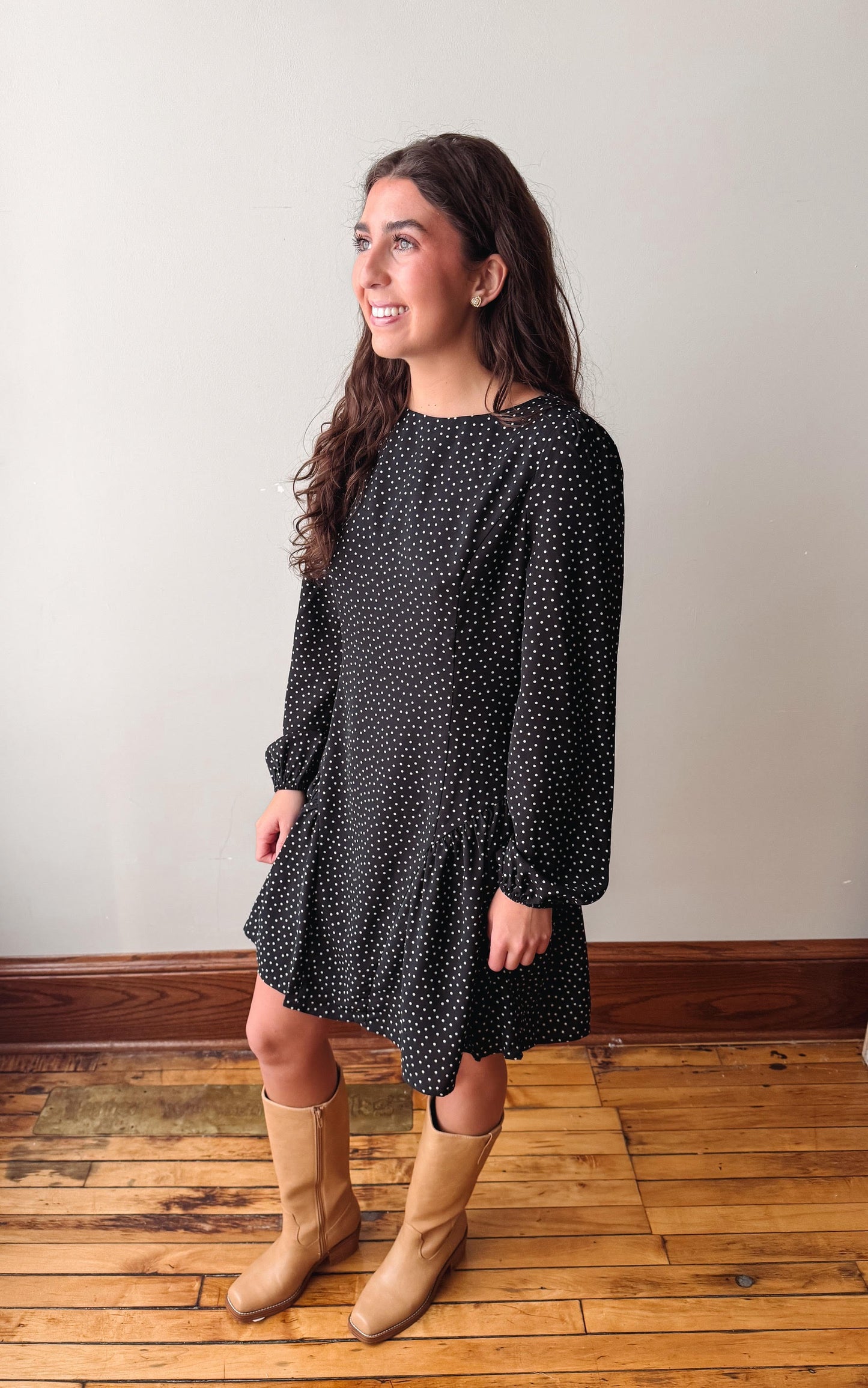 A woman modeling a black long-sleeve dress with a small white polka dot pattern, paired with tan cowboy boots, standing on a wooden floor against a plain wall.