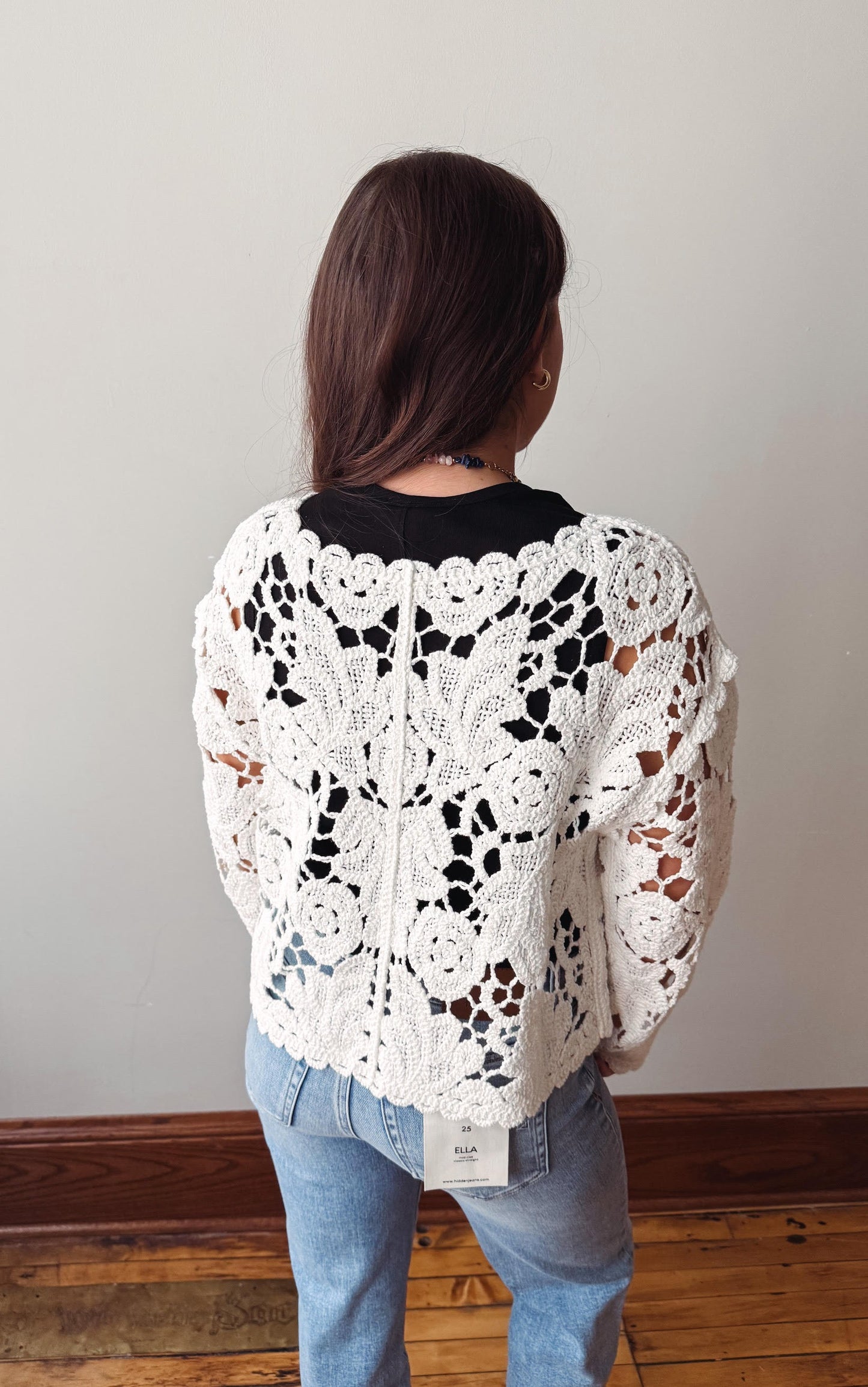 Person wearing a white crochet sweater with black and brown patterns, standing against a plain wall.