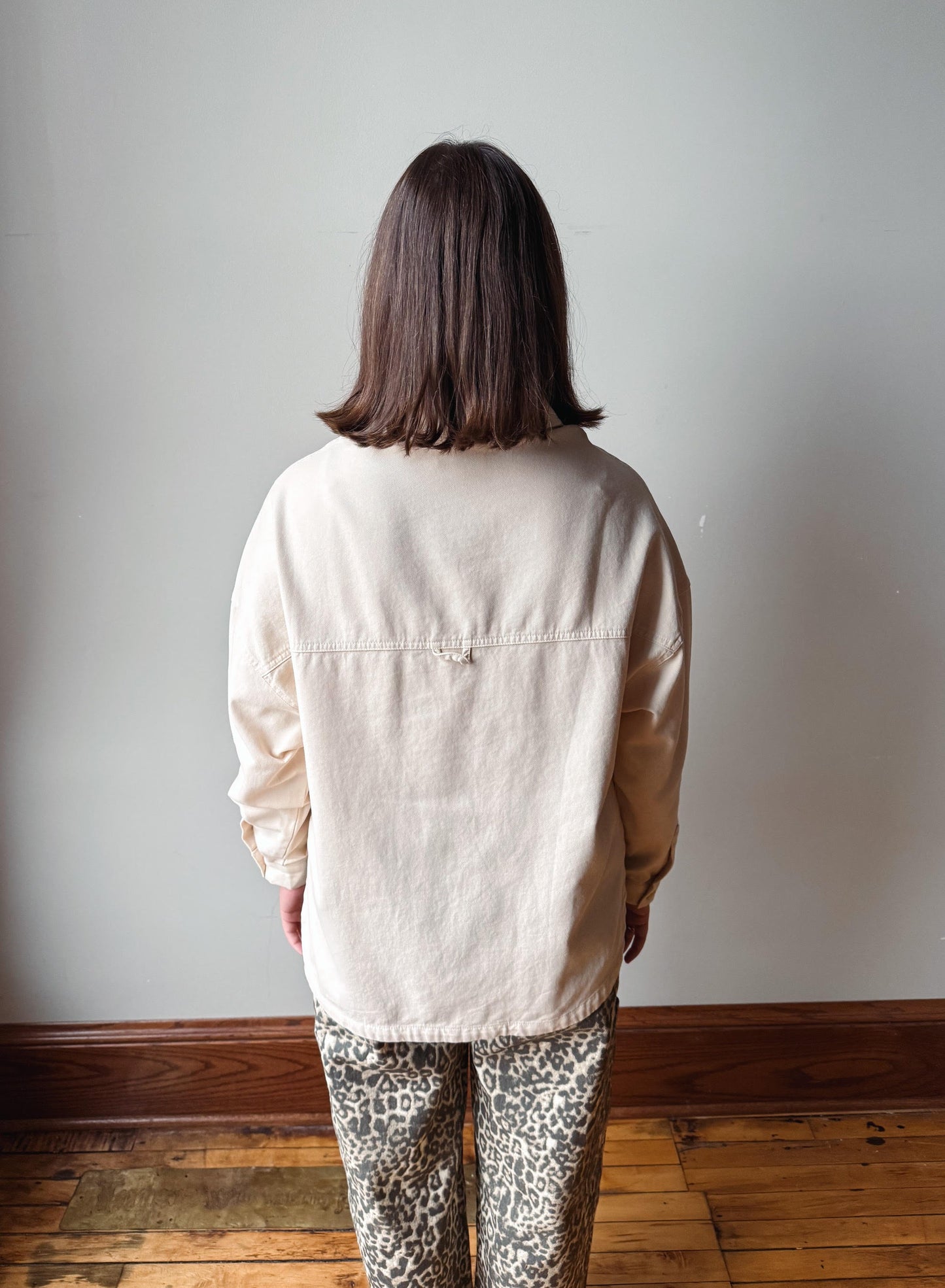 A person viewed from behind wearing a light beige linen button-up shirt and leopard-print pants, standing against a plain light gray wall on a wooden floor.