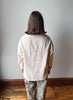 A person viewed from behind wearing a light beige linen button-up shirt and leopard-print pants, standing against a plain light gray wall on a wooden floor.