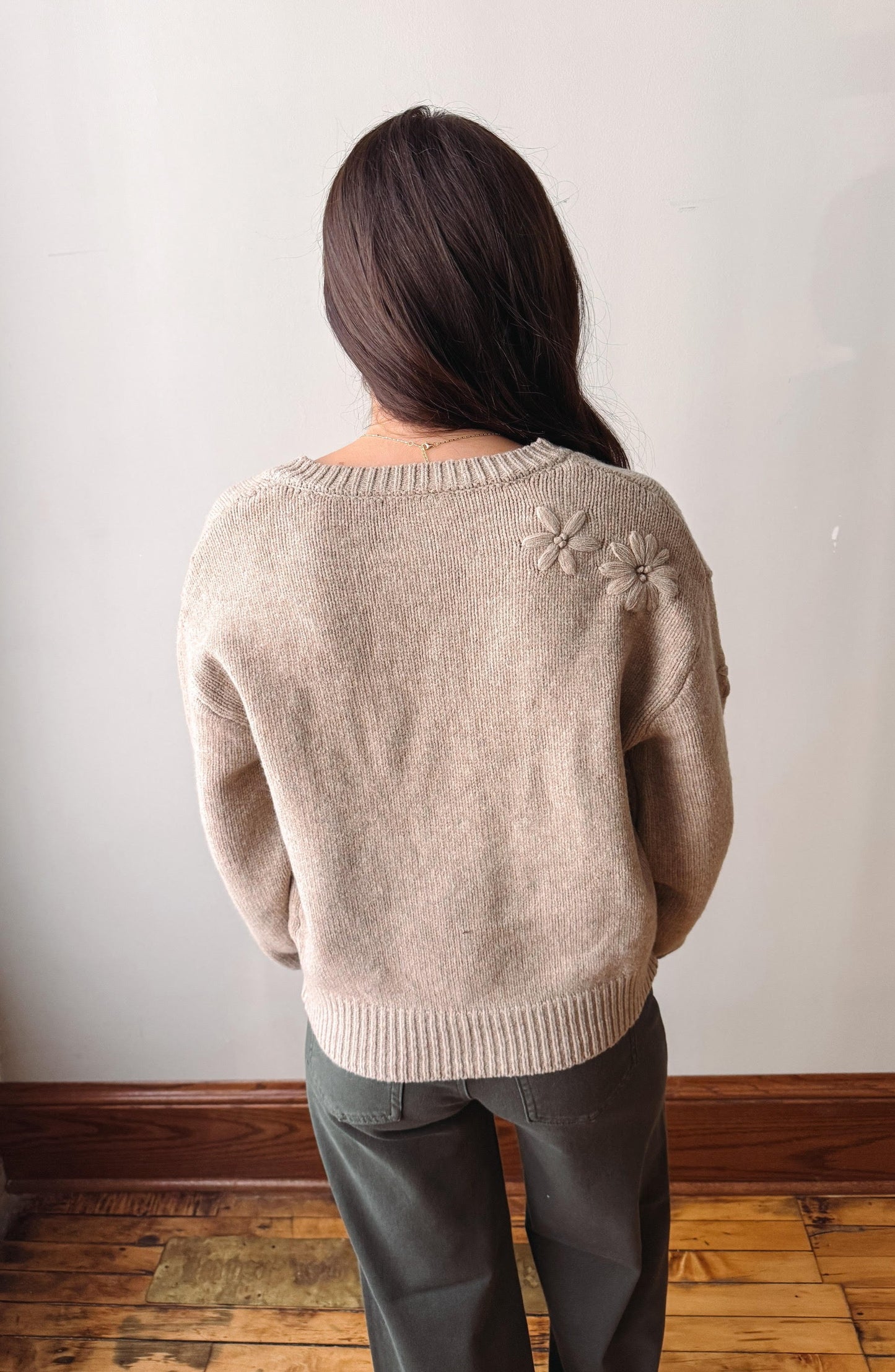 A woman viewed from behind wearing a light beige knit sweater with textured flower details on the upper back and dark gray pants, standing against a plain off-white wall with a wooden floor.