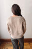 A woman viewed from behind wearing a light beige knit sweater with textured flower details on the upper back and dark gray pants, standing against a plain off-white wall with a wooden floor.
