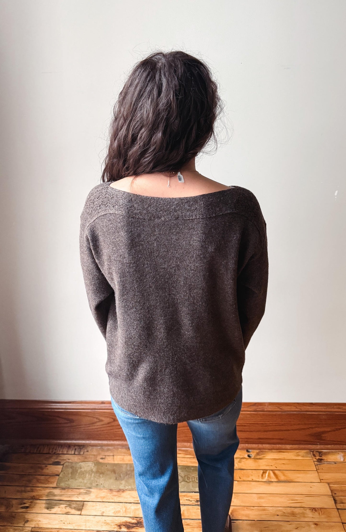A woman viewed from behind wearing a dark brown knit sweater and blue jeans, standing on a wooden floor against a plain off-white wall.