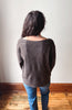 A woman viewed from behind wearing a dark brown knit sweater and blue jeans, standing on a wooden floor against a plain off-white wall.