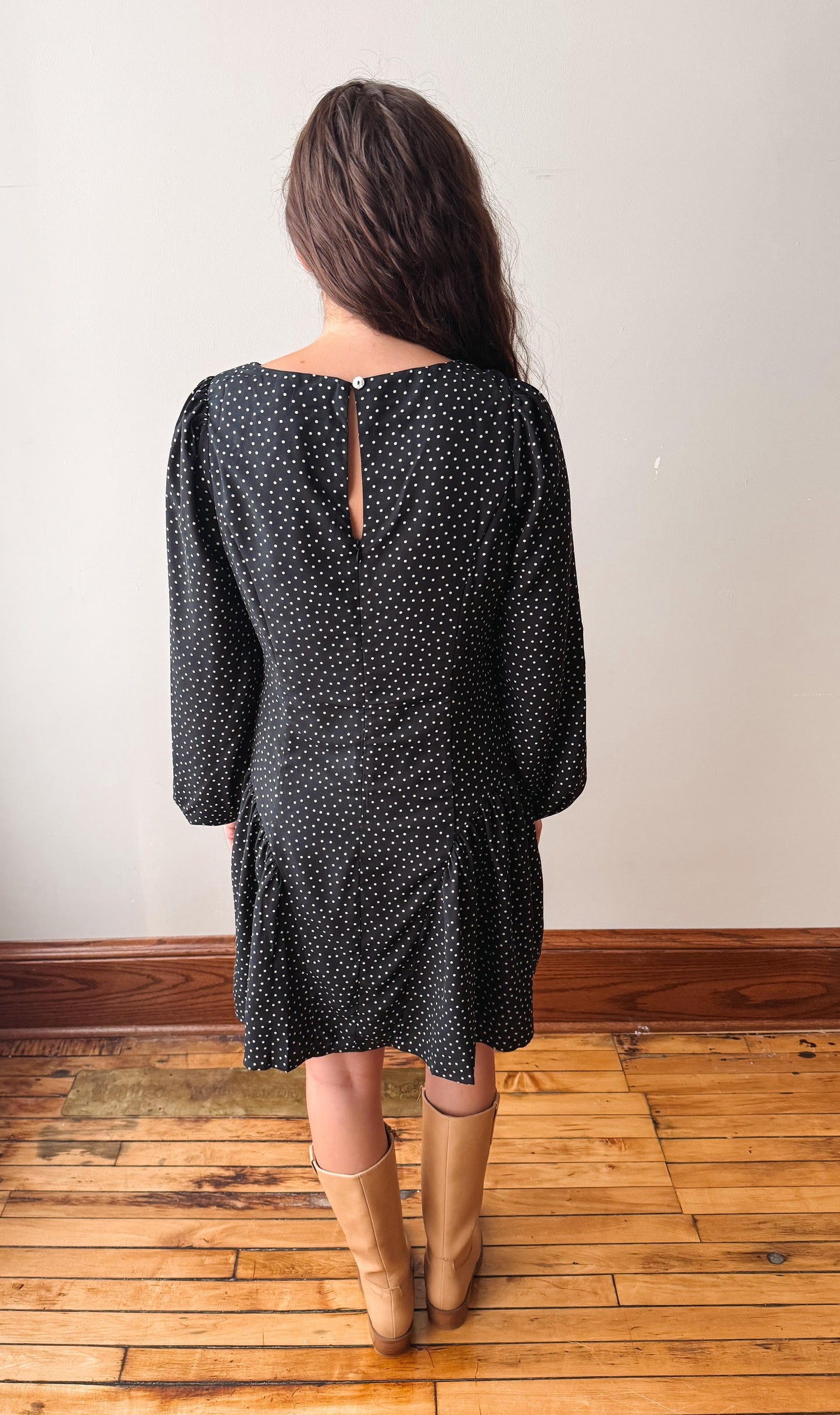 A woman viewed from behind wearing a black long-sleeve dress with a white polka dot pattern and tan knee-high boots, standing on a wooden floor against a plain off-white wall.
