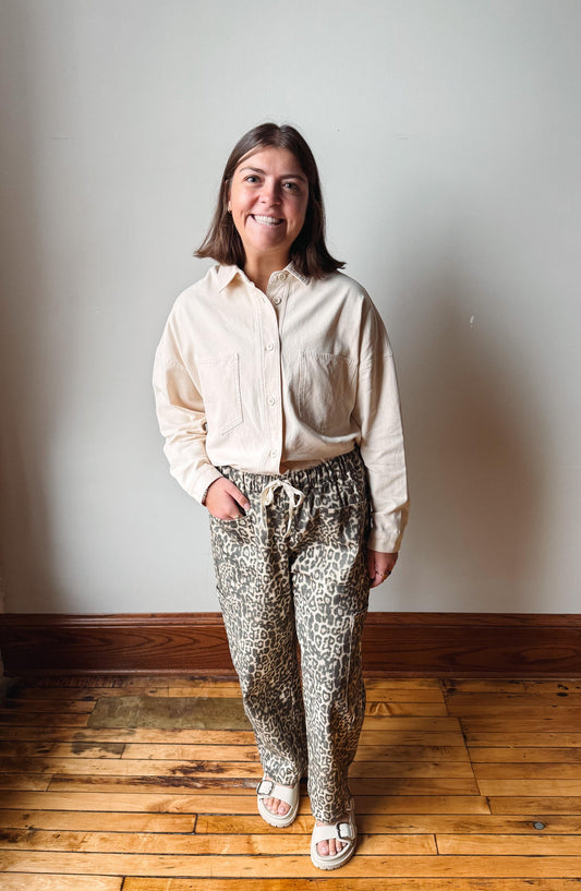 A woman stands in a room with a light gray wall and wooden floor, wearing a cream button-up shirt and leopard-print drawstring pants.