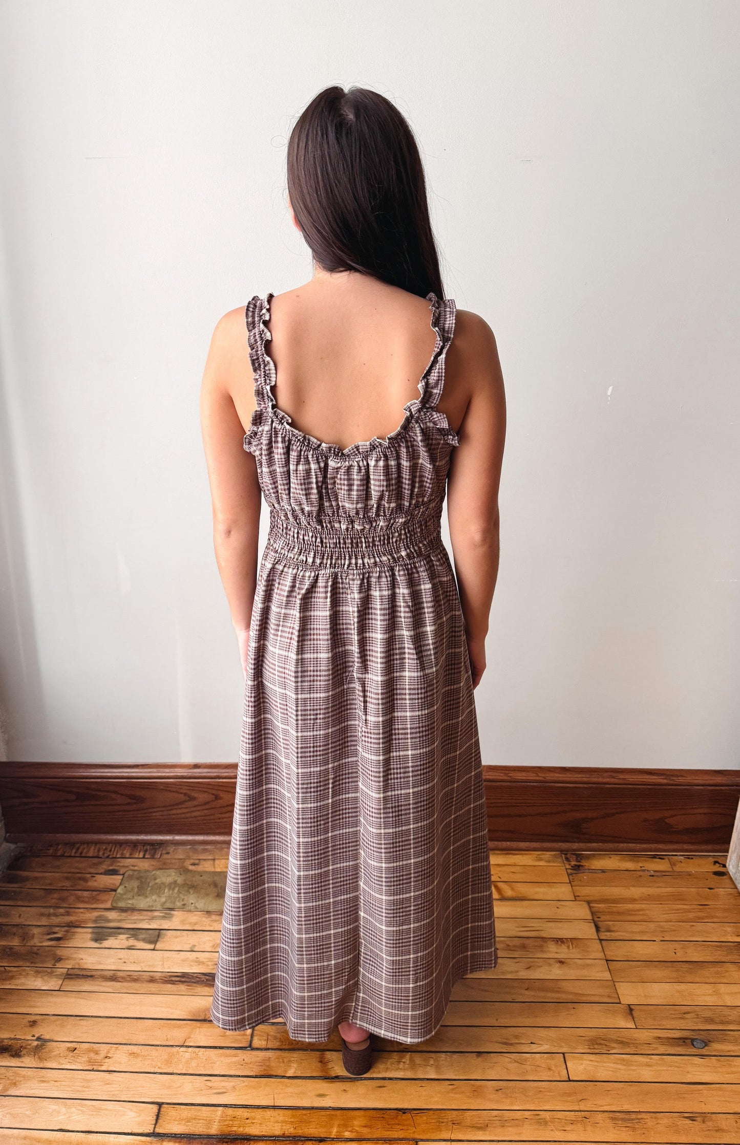 A woman viewed from behind wearing a brown and beige plaid maxi dress with ruffled spaghetti straps and a smocked bodice, standing on a wooden floor against a plain white wall.