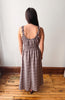 A woman viewed from behind wearing a brown and beige plaid maxi dress with ruffled spaghetti straps and a smocked bodice, standing on a wooden floor against a plain white wall.