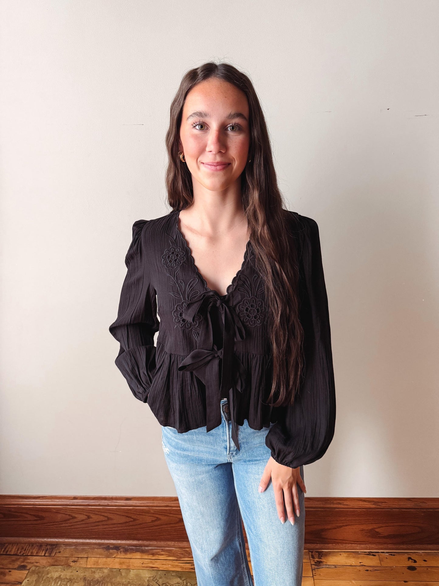 Woman wearing a black blouse and blue jeans standing against a plain wall.