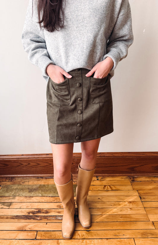 A person wearing a light gray knit sweater, an olive brown mini skirt with front buttons and side pockets, and tan knee-high boots, standing on a wooden floor against a plain wall.