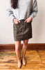 A person wearing a light gray knit sweater, an olive brown mini skirt with front buttons and side pockets, and tan knee-high boots, standing on a wooden floor against a plain wall.