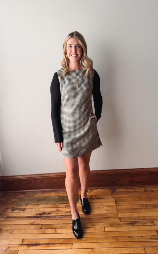 Woman wearing a checkered dress with black sleeves against a plain wall.