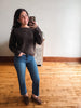 A woman takes a mirror selfie wearing a dark brown fuzzy knit sweater, blue jeans, and brown suede loafers in a room with light wood floors and a white wall.