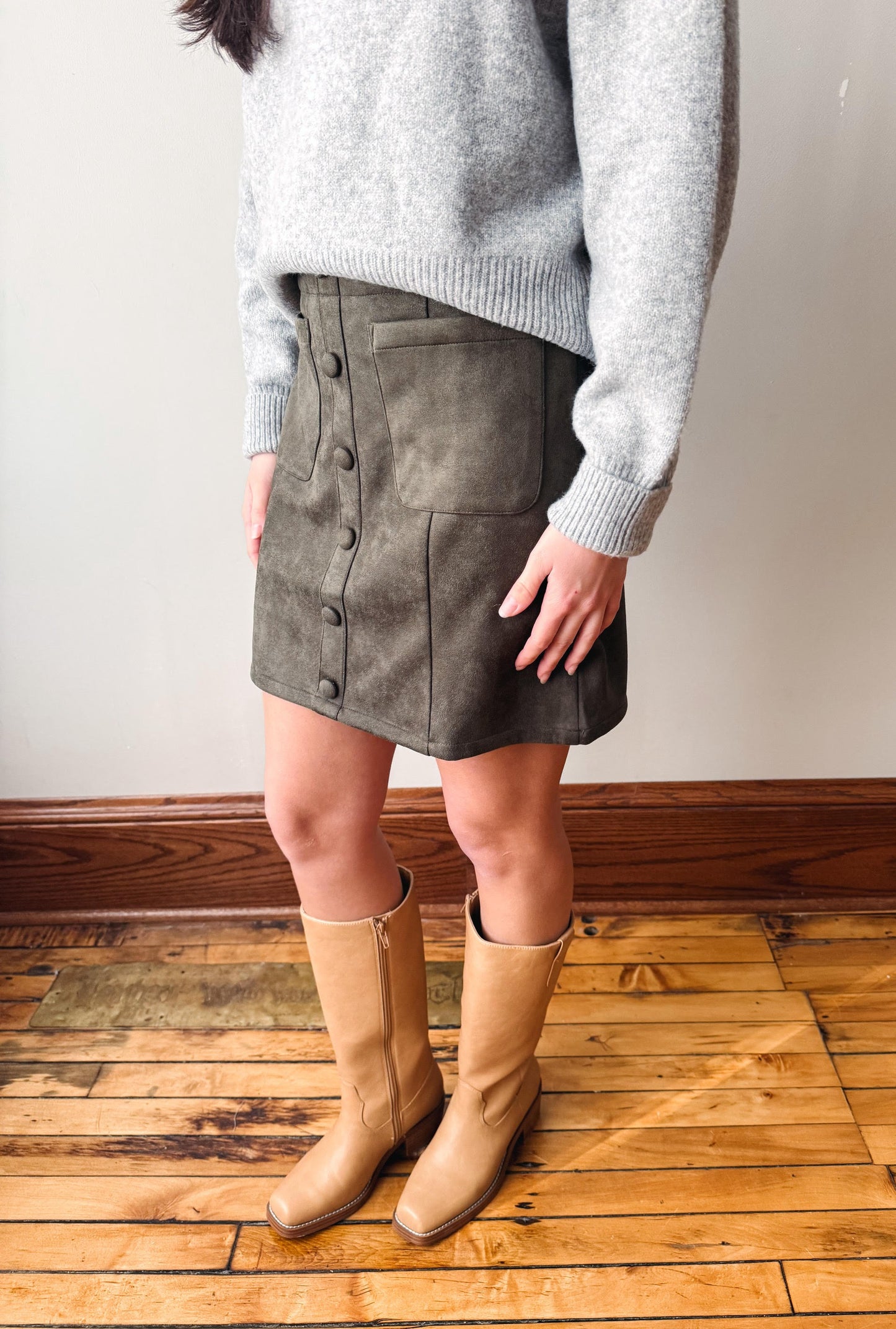 A person wearing a gray knit sweater, a dark gray suede button-front mini skirt, and tan knee-high boots, standing on a wooden floor against a plain wall.