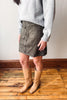 A person wearing a gray knit sweater, a dark gray suede button-front mini skirt, and tan knee-high boots, standing on a wooden floor against a plain wall.