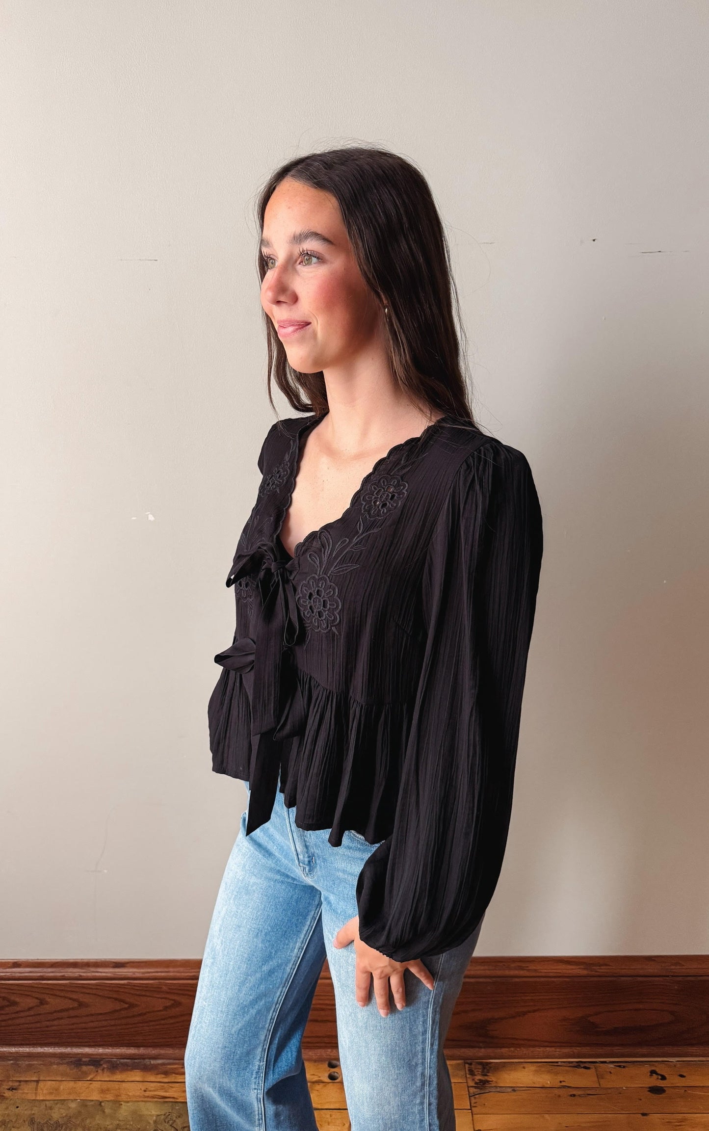 Woman wearing a black blouse with lace details and light blue jeans against a plain wall.