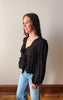 Woman wearing a black blouse with lace details and light blue jeans against a plain wall.