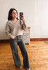 A woman takes a mirror selfie in a room with hardwood floors and a white wall, wearing a beige knit sweater with floral embroidery and dark olive green wide-leg pants.