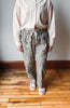 A person wearing loose-fit leopard print drawstring pants, a cream button-up shirt, and white slide sandals, standing on a wooden floor against a plain wall.