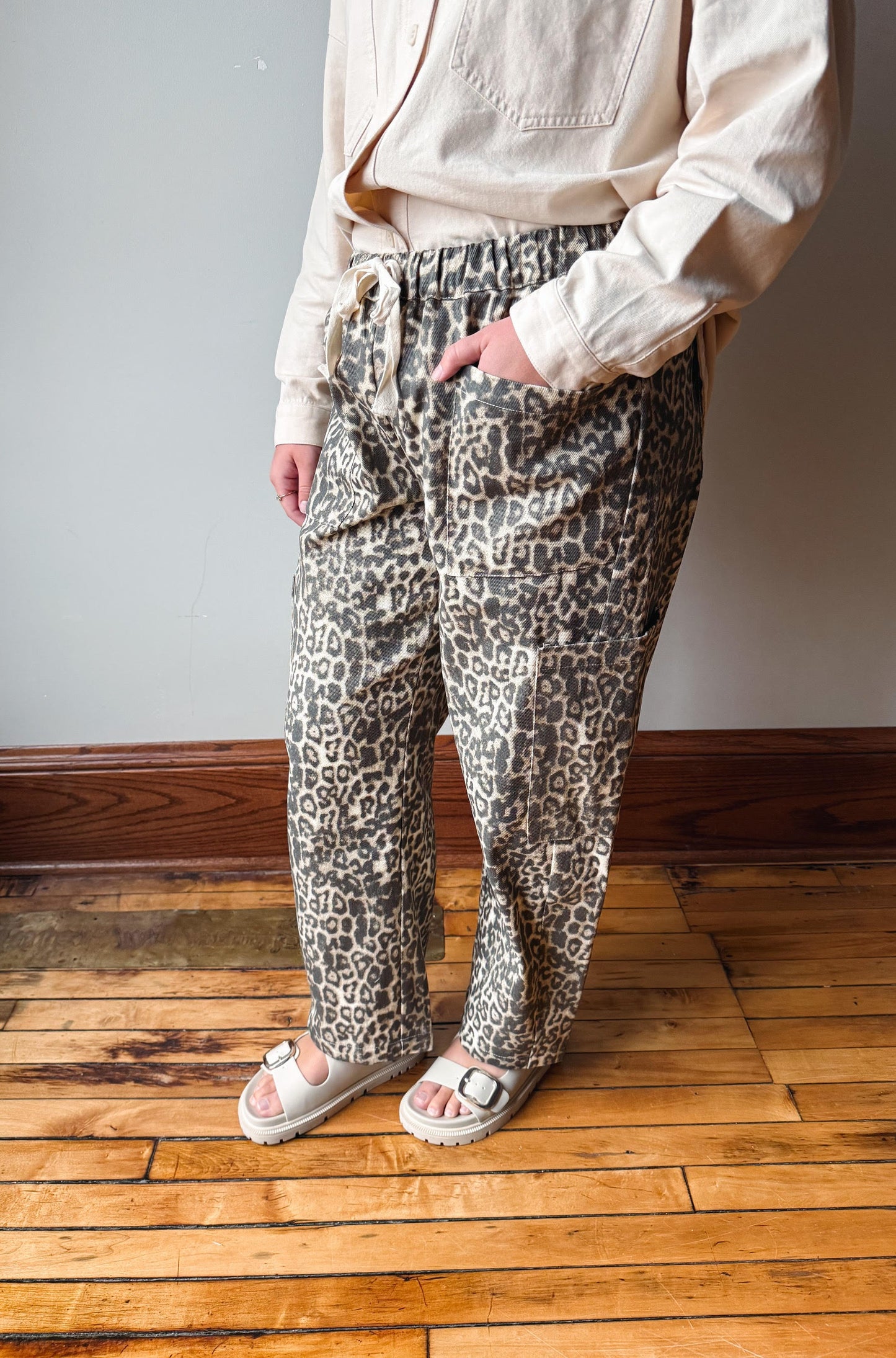 A person wearing loose-fit leopard-print pants with a drawstring waist, paired with a light beige shirt and white slide sandals, standing on a wooden floor against a gray wall.