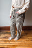 A person wearing loose-fit leopard-print pants with a drawstring waist, paired with a light beige shirt and white slide sandals, standing on a wooden floor against a gray wall.