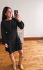 A woman takes a mirror selfie wearing a black long-sleeve mini dress with white polka dots and beige knee-high boots, standing on a wooden floor in front of a white wall.