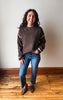 A woman stands indoors wearing a brown knit sweater with cream stripes on the sleeves and cuffs, blue jeans, and brown loafers, against a plain off-white wall and wood floor.