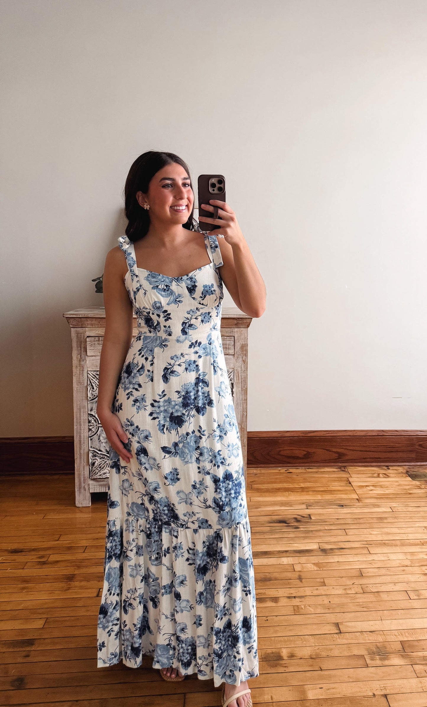 Woman wearing a floral dress taking a mirror selfie in a room with wooden flooring.
