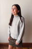 A woman wearing a light gray knit mock neck sweater and a dark brown button-up skirt, standing against a plain beige wall.