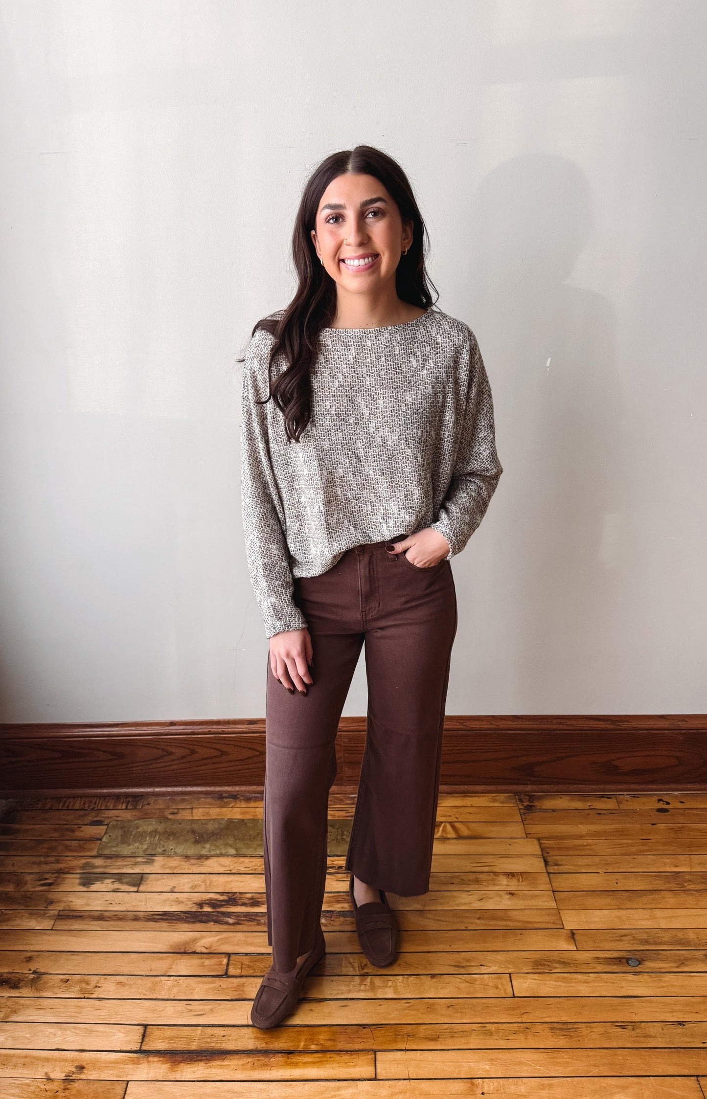 A woman stands in a room with a light gray wall and wooden floor, wearing a textured gray long-sleeve top and dark brown wide-leg pants with matching brown shoes.