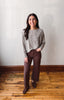 A woman stands in a room with a light gray wall and wooden floor, wearing a textured gray long-sleeve top and dark brown wide-leg pants with matching brown shoes.