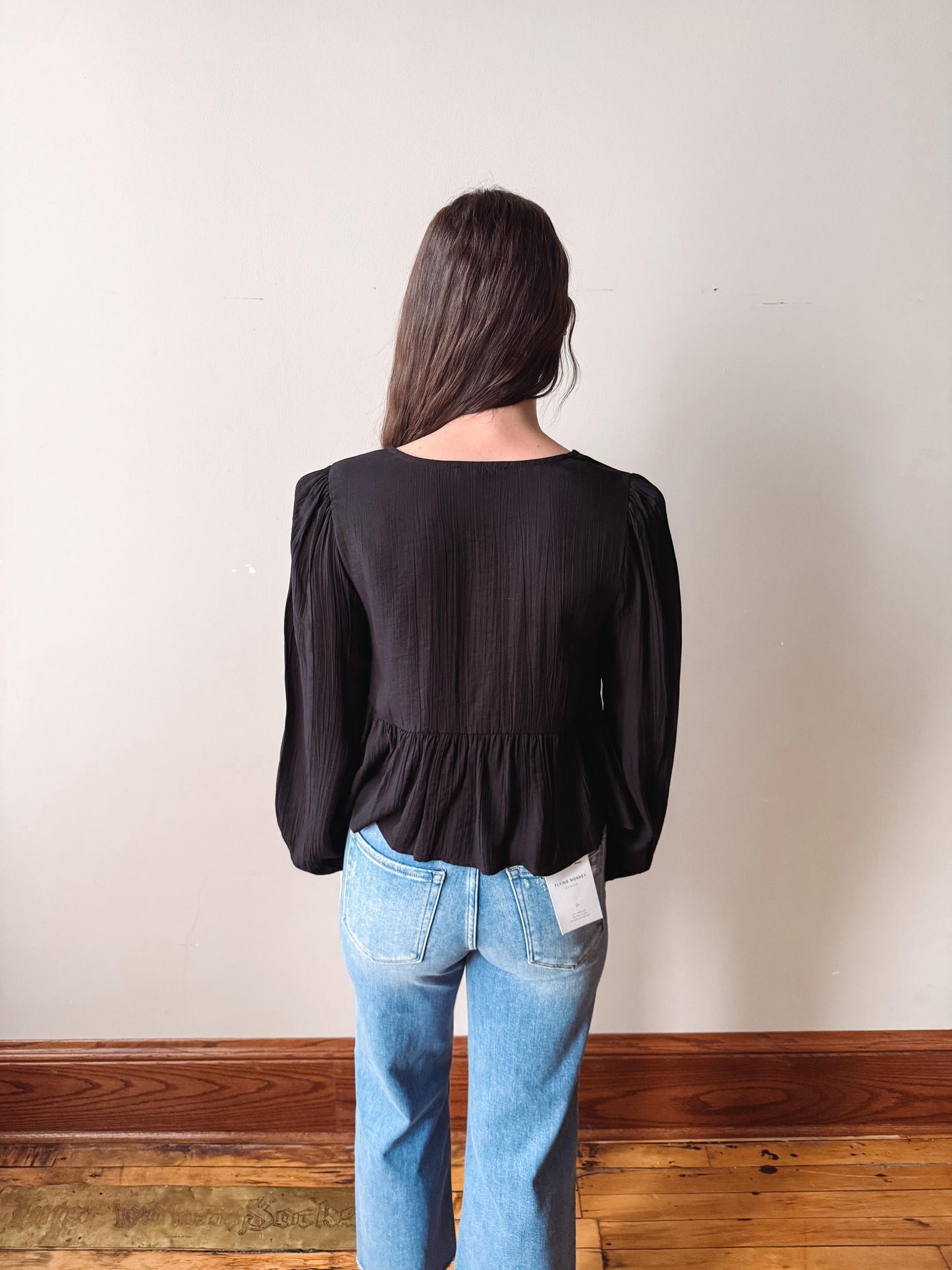 Person wearing a black blouse and blue jeans standing against a plain wall.