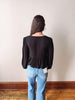 Person wearing a black blouse and blue jeans standing against a plain wall.