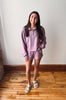 A young woman stands in a room with a white wall and wooden floor, wearing a matching lavender-purple lounge set consisting of a quarter-zip long-sleeve top and shorts, paired with white slide sandals.