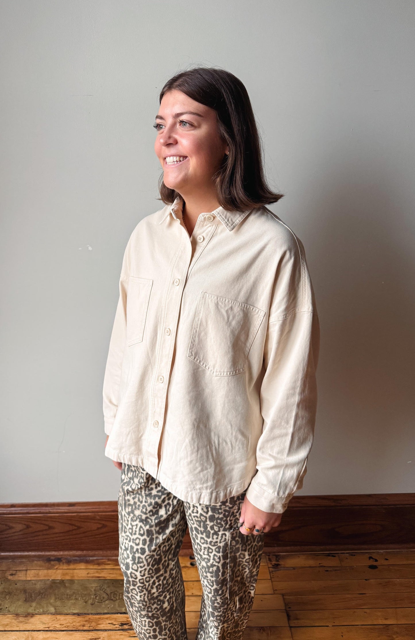 A woman wearing a cream-colored linen button-down shirt and leopard-print pants, standing in a room with a gray wall and wooden floor.