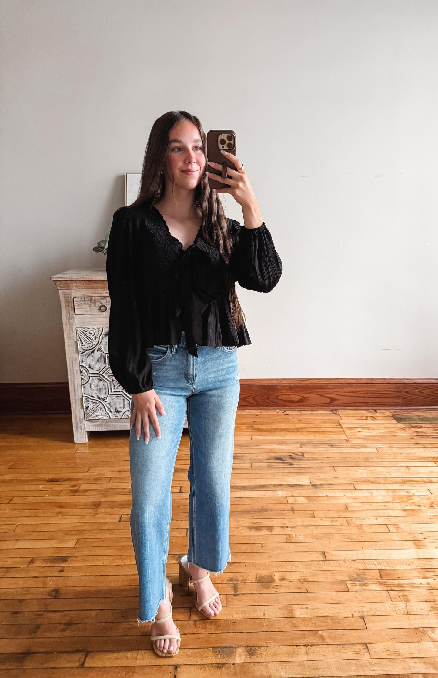 Woman taking a mirror selfie wearing a black blouse and light blue jeans in a room with wooden flooring.
