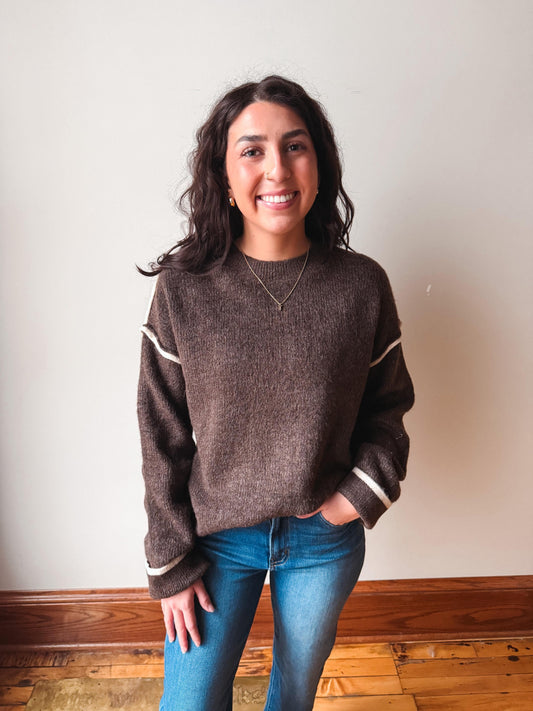 A smiling woman with dark hair stands indoors wearing a brown knit sweater with cream ribbed trim on the sleeves and cuffs, paired with blue jeans, against a plain off-white wall and wood baseboard.