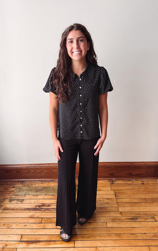 A woman stands in a room with a light beige wall and wooden floor, wearing a black short-sleeve button-up shirt with a small white pattern and black wide-leg pants.