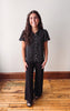 A woman stands in a room with a light beige wall and wooden floor, wearing a black short-sleeve button-up shirt with a small white pattern and black wide-leg pants.