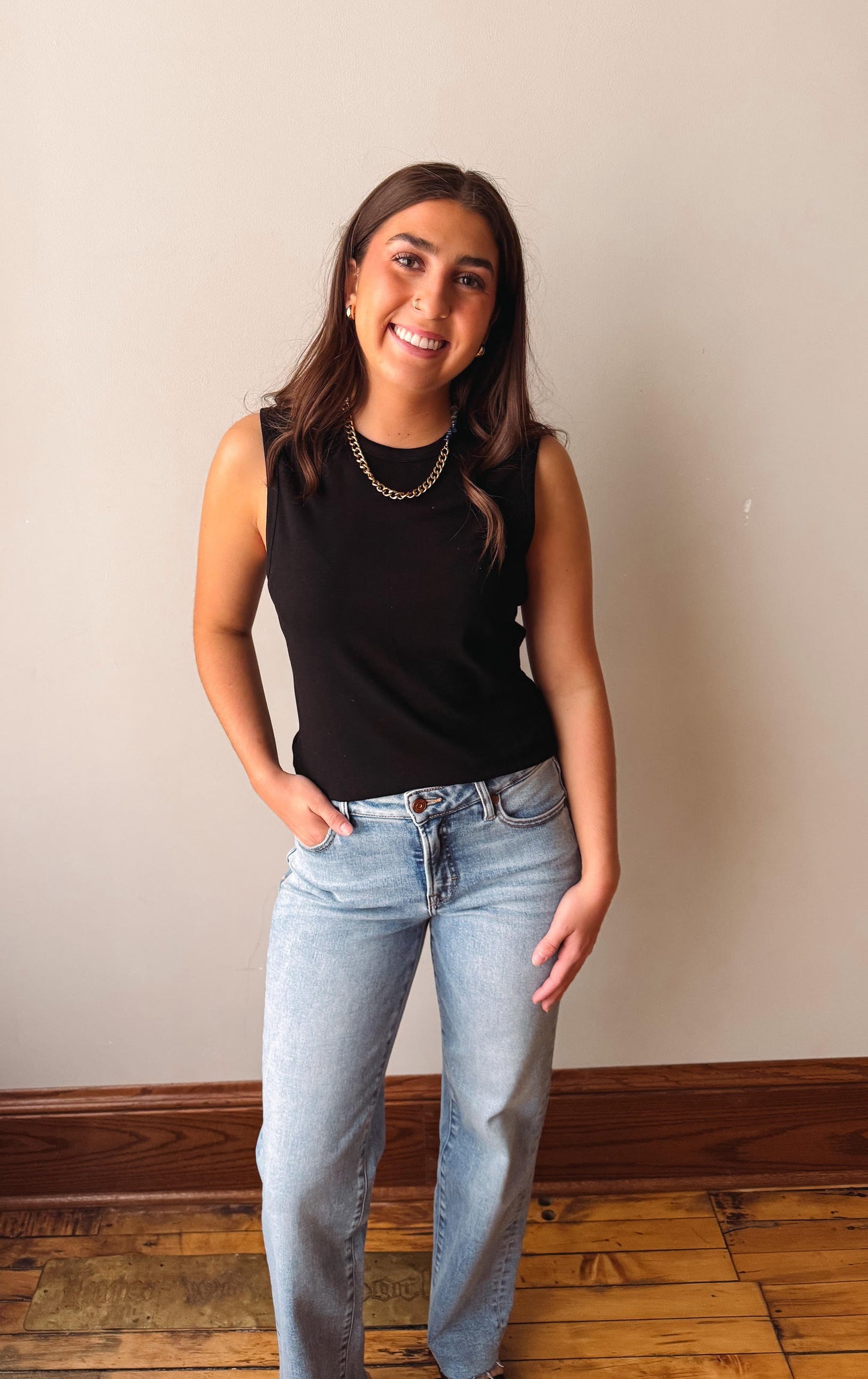 Woman wearing a black sleeveless top and light blue jeans standing against a plain wall.