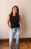 Woman wearing a black sleeveless top and light blue jeans standing against a plain wall.