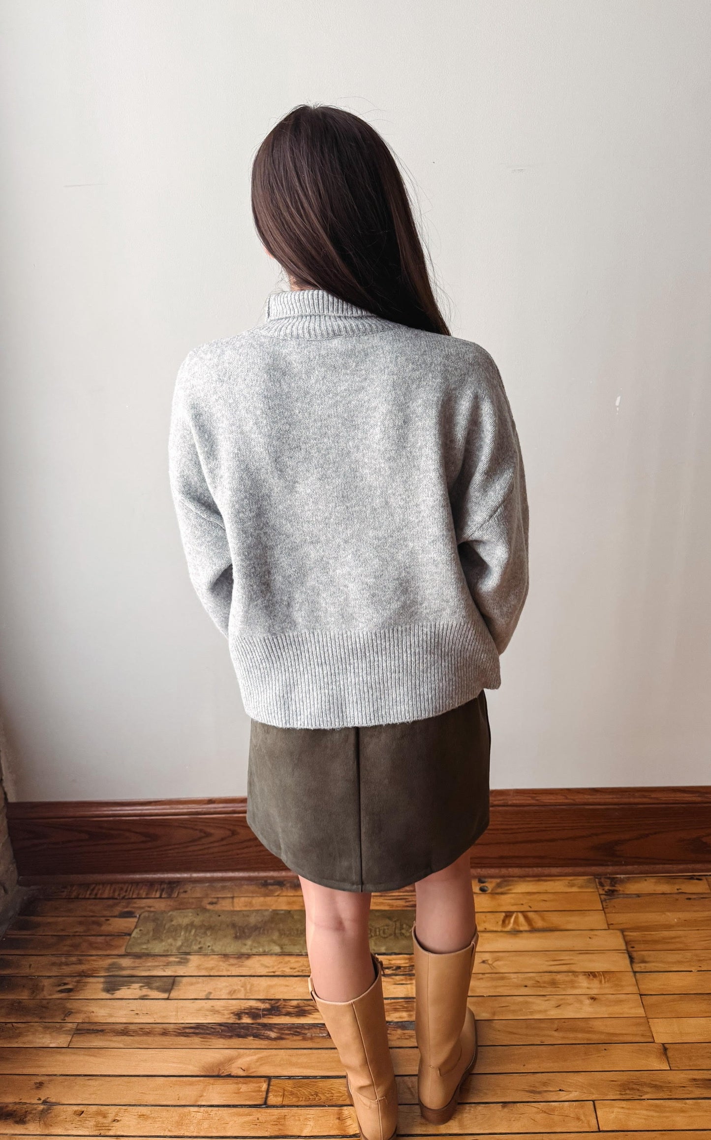 A woman viewed from behind wearing a light gray knit sweater, a dark brown mini skirt, and tan knee-high boots, standing on a wooden floor against a plain off-white wall.