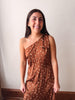 A woman smiling and wearing a brown one-shoulder dress with white polka dots, standing against a plain off-white wall.